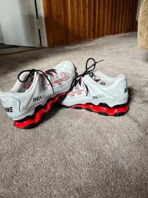 Nike Reax sneakers - Nike Reax sneakers i vitt med röda och svarta detaljer. Skorna har en sportig design med svart sula och röda inslag i mellansulan. Ovandelen är i syntetmaterial med svarta skosnören och tydliga Nike-loggor på sidorna och hälen.