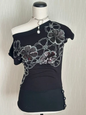 Svart stickad topp med paljettblommor - Svart stickad topp med asymmetrisk offshoulder-design och stora blommor i grått och silver, dekorerade med paljetter. 