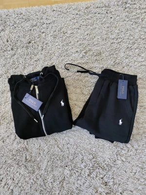 Svart tracksuit från Polo Ralph Lauren - Snyggt svart tracksuit från Polo Ralph Lauren med dragkedjetröja och matchande byxor. Setet har den klassiska vita loggan broderad på både tröjan och byxan. Tröjan har huva och fickor, byxorna har dragsko och resår i midjan.