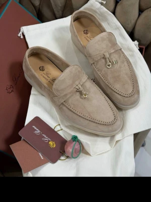 Beige loafers från Loro Piana - Snygga beige loafers i mocka från Loro Piana med dekorativt guldfärgat spänne framtill. Skorna har klassisk design med rundad tå och diskret logga på hälen. Perfekta för en stilren och trendig look.