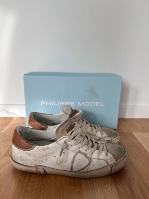 Vita Philippe Model sneakers med bruna detaljer - Box, Och kvitto medföljer, om det behlvs tvätt innan jag skickar dom så är det bara att fråga. Liten defekt vid sulan men det går att använda som vanligt. Snygga vita sneakers från Philippe Model med bruna detaljer vita snören och diskret logga på sidan. Tillverkade i läder med gummisula, perfekta för en clean och stilren look.