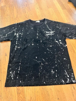 Svart t-shirt med vitt stänkmönster WPZ - Cool svart t-shirt från WPZ med vitt stänkmönster över hela tröjan och tryck på bröstet. Klassisk rund hals och korta ärmar. Perfekt för dig som gillar streetwear och vill sticka ut lite extra.