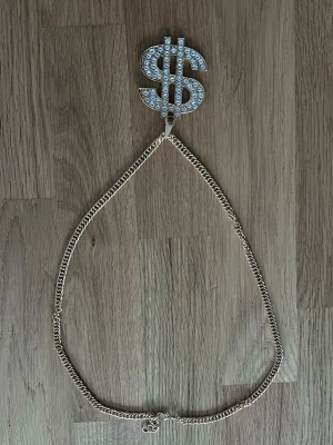 Guldhalsband med blingad dollartecken - Snyggt halsband i guldfärgad metall med en stor dollar-symbol som är täckt av glittrande pärlor. Kedjan är klassisk och har ett coolt statement-hänge som verkligen sticker ut. Perfekt för dig som vill ha något unikt och vågat.