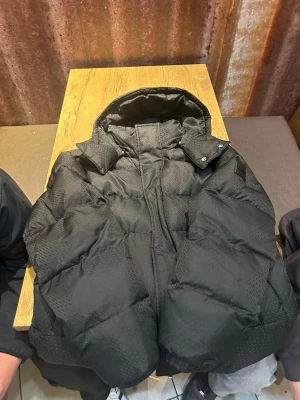 Svart dunjacka från BOSS med huva - Snygg svart dunjacka från BOSS med diskret rutmönster i tyget. Jackan har en stor huva och dragkedja framtill. Perfekt för kalla vinterdagar och ger en modern look med sin stilrena design.