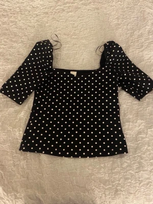 Svart prickig blus med puffärm - Trendig svart blus med vita prickar och puffiga halvlånga ärmar. Fyrkantig ringning och knytband vid axlarna ger en söt och modern look. Perfekt att styla med jeans eller kjol för en cool outfit.