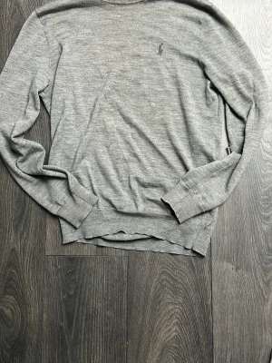 Grå tröja från Ralph Lauren - Klassisk grå tröja från Ralph Lauren med diskret broderad logga på bröstet. Tröjan har rund halsringning, långa ärmar och ribbade muddar. Perfekt för en stilren och enkel look. Tröjan har hål vid armbågarna där av de låga priset. Förändrar itne användningen av tröjan.