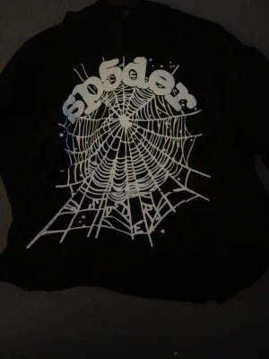 Svart hoodie från SP5DER med tryck - Svart hoodie från SP5DER med stort vitt spindelnäts-tryck och texten 'sp5der' på bröstet. Oversized streetwear-stil med huva och klassisk passform. Perfekt för dig som gillar unika och grafiska plagg.