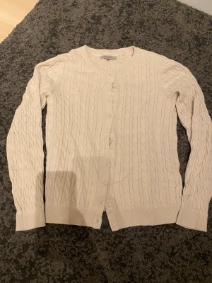 Beige kabelstickad kofta - Kabelstickad beige kofta med rund halsringning och knäppning framtill. Tillverkad i bomull och har långa ärmar samt ribbade muddar. Perfekt för lager-på-lager och enkel att matcha med olika outfits.