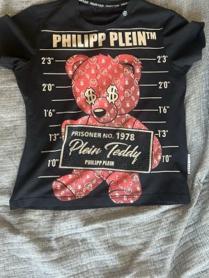 Svart PP t-shirt med teddy - Cool svart t-shirt med stort tryck av en röd teddy med dollartecken på framsidan. Baksidan har ett rött dollartecken. Perfekt statement-plagg för dig som gillar streetwear. Bra kvalité och använd fåtal gånger! 😌