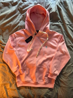 Rosa hoodie från Polo Ralph Lauren - Snygg rosa hoodie från Polo Ralph Lauren med klassisk huva och vit dragsko. Har en broderad blå logga på bröstet och en stor magficka. Perfekt för en chill och trendig look. Mjuk bomullskvalitet och ribbade muddar vid ärmslut och nederkant.