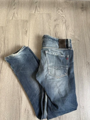 Blå slim fit jeans från Replay - Säljer ett par blå slim fit jeans från Replay med snygga slitningar och tvättade detaljer. Klassisk femficksmodell med låg midja och dragkedja. Perfekta för en avslappnad och trendig look.