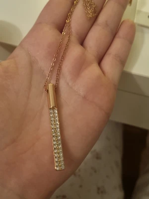 Guldigt halsband med stavhänge - Snyggt halsband i guldton med ett avlångt stavformat hänge täckt av glittrande stenar. Kedjan är tunn och elegant, perfekt för att ge lite extra bling till din outfit. Hänget har en modern och stilren design som sticker ut.