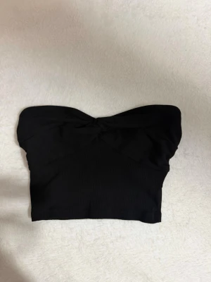 Svart bandeau tubtopp - Snygg svart bandeau tubtopp med twistad detalj framtill. Toppen är axelbandslös och har en enkel, stilren design som passar perfekt till många olika outfits. Samt finns det gummi som gör att tröjan inte åker ner och visar inte för mycket. 