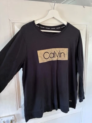 Svart tröja från Calvin Klein - Svart långärmad sweatshirt från Calvin Klein med rund halsringning och ett stort guldfärgat tryck med texten 'Calvin' på bröstet. Mjuk bomullskvalitet, perfekt för en avslappnad och trendig look. Passar snarare M.