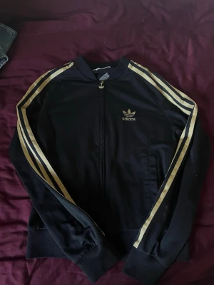Svart Adidas jacka med guldiga ränder - Svart jacka från Adidas med klassiska guldiga ränder längs ärmarna och broderad logga på bröstet. Storlek 38