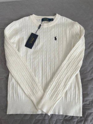 Vit kabelstickad tröja Polo Ralph Lauren - Vit kabelstickad tröja från Polo Ralph Lauren med rund halsringning och klassisk broderad logga på bröstet. Tröjan har ribbade muddar och lång ärm, perfekt för en clean och stilren look.
