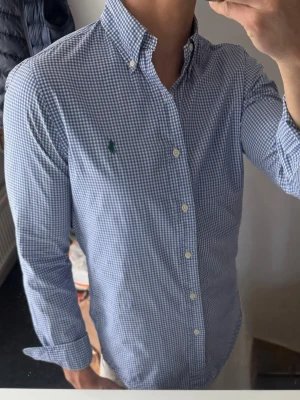 Blårutig skjorta från Polo Ralph Lauren - Klassisk blårutig skjorta från Polo Ralph Lauren med button-down krage och lång ärm. Skjortan har små vita och blå rutor samt den ikoniska gröna logotypen broderad på bröstet. Tillverkad i bomull för en skön och luftig känsla. Pytteliten reva mitt på, men syns knappt.