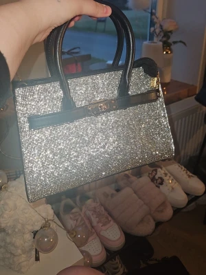 Glittrig silver handväska med svarta detaljer - Snygg handväska i glittrigt silver med svarta handtag och dekorativt spänne framtill. Väskan har en rektangulär form och är perfekt för dig som vill sticka ut. Materialet är täckt av små glittrande stenar och detaljerna är i svart konstläder. Nypris 499kr helt ny köpt I Danmark 