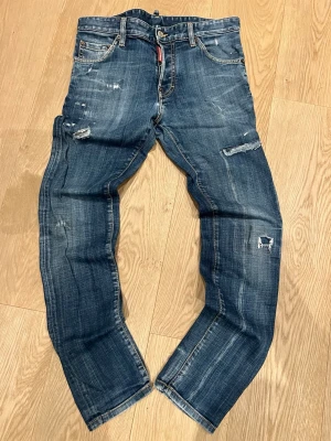 Blå slitna jeans från  - Säljer ett par blå jeans från  Dsquared2 med slitningar och distressed detaljer på både fram- och baksida. Klassisk femficksmodell med raka ben och snygg tvätt. Jeansen har en röd Dsquared2-tag vid fickan och är tillverkade i bomull.