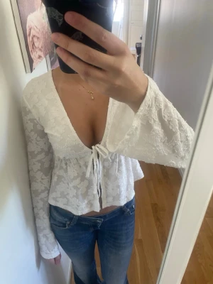 Vit blus med knyt från Newyorker  - Superfin vit blus från Newyorker med v-ringning och knyt framtill. Blusen har långa ärmar och är täckt av ett romantiskt spetsmönster. Den är lätt utsvängd nedtill och har en feminin, luftig känsla. Perfekt att styla med jeans eller kjol.