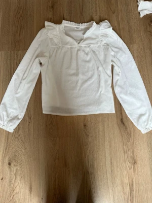 Vit blus med volang från Kappahl - Snygg vit blus från Kappahl med långa ärmar och volangdetaljer vid axlar och halsringning. Blusen har en lätt rynkad struktur i tyget och en rak passform. Perfekt för dig som gillar en romantisk och trendig stil.