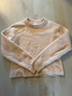 Ljusrosa stickad tröja från H&M - Mysig ljusrosa stickad tröja från H&M med rund halsringning och långa ärmar. Tröjan har en fluffig, mjuk struktur och ribbade muddar vid ärmslut och nederkant. 