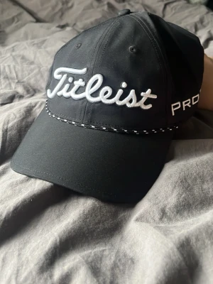 Svart Titleist keps med brodyr - Snygg svart keps från Titleist med vit broderad logga framtill och på baksidan. Kepsen har böjd skärm, justerbar rem bak och detaljer i vitt längs skärmen. Perfekt för dig som gillar sportig stil och golf.