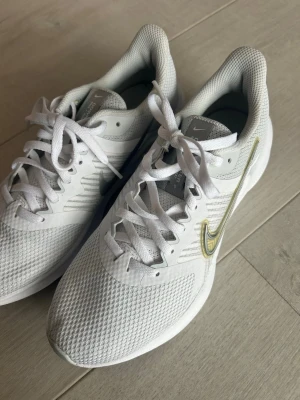 Vita Nike löparskor med gulddetalj - Snygga vita Nike sneakers med meshmaterial för bra ventilation och en guldfärgad Swoosh på sidan. Skorna har klassisk låg profil, vita snören och en bekväm, vadderad sula som passar perfekt för träning eller promenader.
