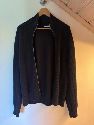 Mörkblå stickad ziptröja - Snygg svart stickad ziptröja med hög krage och ribbade muddar. Tröjan har en rak passform och är tillverkad i ett mjukt, tjockare material som passar perfekt till kyligare dagar.