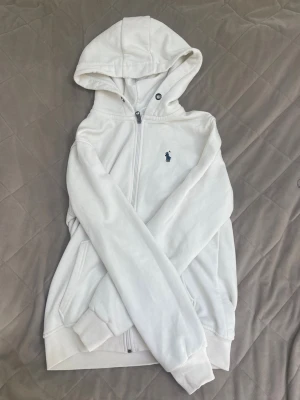 Vit hoodie från Polo Ralph Lauren - Snygg vit hoodie från Polo Ralph Lauren med dragkedja framtill och klassisk blå logga på bröstet. Hoodien har huva, ribbade muddar och fickor på sidorna. Perfekt för en clean och stilren look. Materialet är mjukt och bekvämt, troligen bomullsblandning.