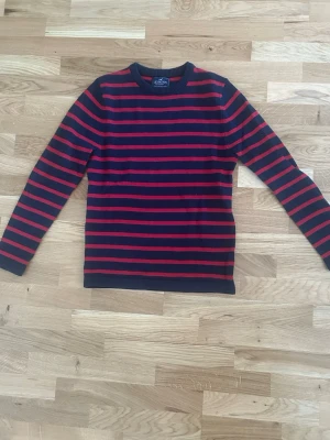 Randig stickad tröja från Lexington - Mörkblå stickad tröja från Lexington med röda horisontella ränder. Tröjan har rund halsringning och ribbstickade muddar vid ärmslut och nederkant. Perfekt för dig som gillar klassisk och stilren design.