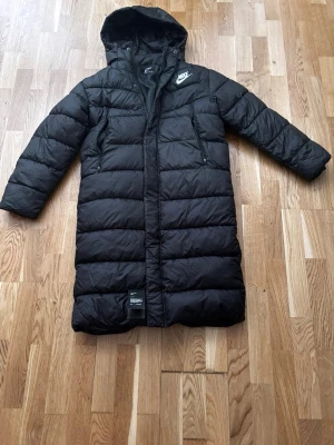 Lång Jacka - Svart lång dunjacka från Nike med huva och dragkedja framtill. Jackan har två sidofickor med dragkedja och en vit Nike-logga på bröstet. Perfekt för kalla vinterdagar och har en stilren, sportig look.