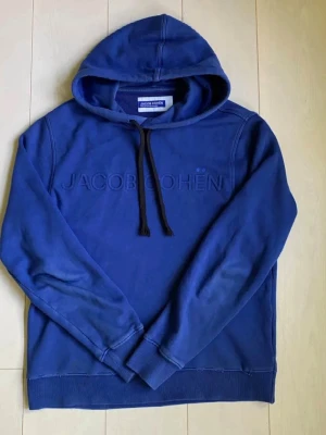 Blå hoodie från Jacob Cohën - Snygg blå hoodie från Jacob Cohën med broderad logga på bröstet och svarta dragsnören i huvan. Hoodien har ribbade muddar vid ärmslut och nederkant. Perfekt för en avslappnad och trendig stil. Nypris 5k