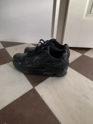 Svarta Nike Air Max sneakers - Snygga helsvarta Nike Air Max sneakers med klassisk siluett och synlig luftdämpning i sulan. Skorna har en låg profil, snörning och är tillverkade i en mix av läder och syntetmaterial. Perfekta för dig som gillar en clean och sportig stil.