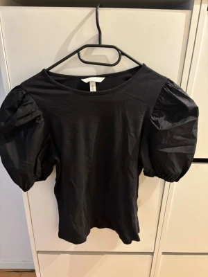 Svart topp med puffärmar från H&M - Svart topp från H&M med trendiga puffärmar och rund halsringning. Toppens material är mjukt och stretchigt, vilket gör den bekväm att bära. Perfekt för dig som vill ha en stilren men ändå iögonfallande look.