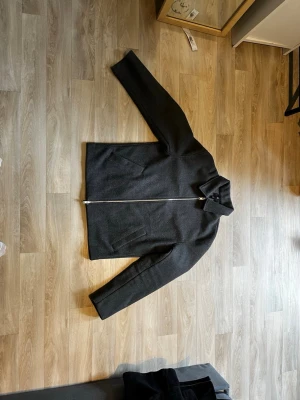 Mörkgrå ulljacka med dragkedja. V i kan förhandla  - Stilren mörkgrå jacka i ull med rak passform och klassisk krage. Jackan har hel dragkedja framtill och två snedställda fickor. Perfekt för dig som gillar minimalistisk och clean stil.