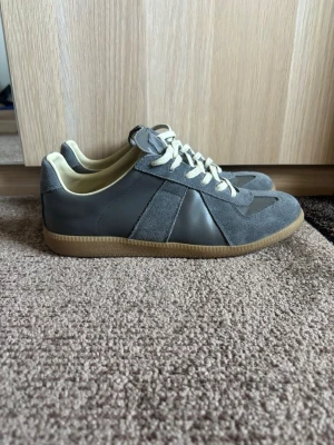 Grå maison margiela gats - Stilrena grå sneakers med ovandel i både mocka och läder, vita snören och beige innersida. Skorna har en klassisk gummisula i ljusbrunt och en låg silhuett som passar till många olika outfits. Perfekta för dig som gillar en clean och tidlös look.