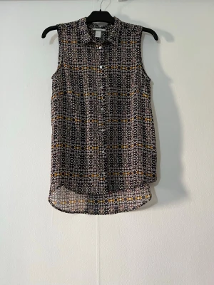 Mönstrad ärmlös blus från H&M - Snygg ärmlös blus från H&M med svart, vit och gul geometrisk mönstring. Blusen har krage, knäppning framtill och är tillverkad i ett lätt och luftigt material. Perfekt till varma dagar och enkel att styla med jeans eller kjol.