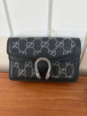 Gucci Dionysus svart handväska - Snygg svart handväska från Gucci med klassiskt GG-mönster i vitt och ikoniskt silverspänne i form av ett hästskohuvud. Väskan är tillverkad i canvas med läderdetaljer och har en kompakt, rektangulär form. Perfekt för dig som gillar lyxiga accessoarer.
