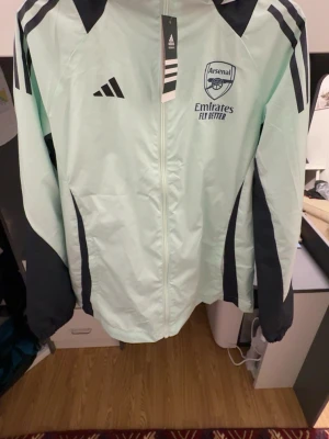 Arsenal vindjacka från Adidas - Snygg mintgrön vindjacka från Adidas med Arsenal-logga och 'Emirates Fly Better' tryck på bröstet. Jackan har svarta detaljer på ärmar och axlar, dragkedja framtill och huva. Perfekt för dig som vill visa ditt stöd för Arsenal och hålla stilen.
