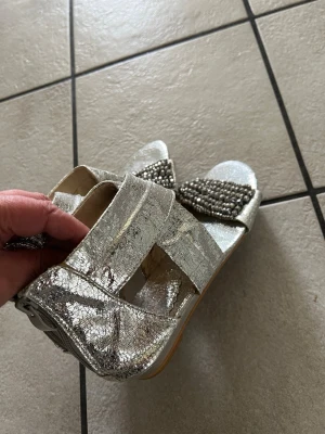 Silvriga sandaler med pärldetaljer storlek 37 - Snygga sandaler i metallic-silver med breda remmar och öppen tå. Framtill pryds de av en rad med glittrande pärlor som ger extra bling. Sandalerna har dragkedja bak och är tillverkade i ett glansigt syntetmaterial. Perfekta för att sticka ut i mängden.