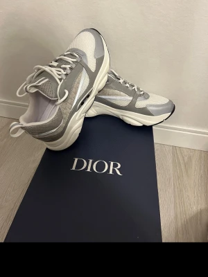 Gråvita sneakers dioor - Snygga sneakers från Dioor i vitt och grått med meshpaneler och detaljer i läder. Skorna har en chunky sula och klassisk snörning. Perfekta för dig som gillar exklusiva och trendiga sneakers med modern design.