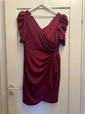 Vinröd omlottklänning med puffärm - Vinröd kort omlottklänning med puffärmar och draperad detalj i midjan. Klänningen har v-ringning både fram och bak samt korta ärmar. Perfekt för dig som vill sticka ut med en elegant och trendig look.