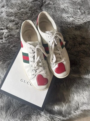Gucci vita sneakers med hjärtdetalj - Unika vita sneakers från Gucci i läder med röda hjärtan på tårna och klassiska gröna och röda ränder på sidorna. Skorna har vita snören och låg profil. Perfekta för dig som vill sticka ut med en cool och lekfull detalj. 