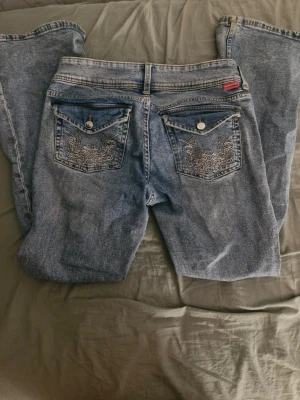 Ljusblåa bootcut jeans med fin design på fickorna - Ganska lågmidjade bootcut jeans, originellt från H&M. Lite slitna längs ned vid fötterna men annars inga skador. Pris kan diskuteras!