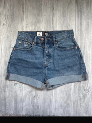Blå denimshorts från lager 157 XS NWT - Säljer ett par klassiska blå denimshorts från lager 157 med hög midja och uppvikta benslut. Shortsen har fem fickor, bälteshällor och stängs med knappar framtill. Perfekta till sommaren och lätta att matcha med allt.