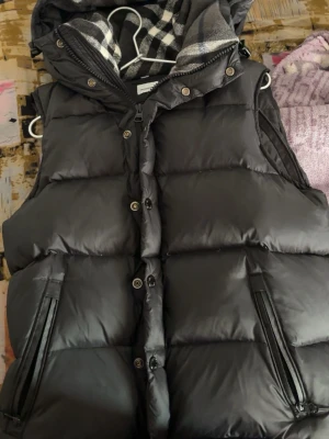 Svart burberry väst  - Snygg svart burberry väst med dragkedja och knappar framtill, två fickor med dragkedja och avtagbar huva med rutigt foder. Perfekt för lager-på-lager och håller dig varm under kyliga dagar.