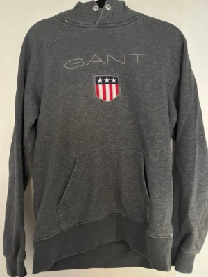 Grå hoodie från GANT med brodyr - Grå hoodie från GANT med broderad logga och amerikanskt inspirerat emblem på bröstet. Klassisk känguruficka framtill, långa ärmar och huva med snörningsdetalj. Perfekt för en chill och stilren look.
