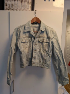Ljusblå jeansjacka från Åhlens  - Snygg ljusblå jeansjacka från Åhlena i storlek S. Klassisk modell med två bröstfickor med lock och knappar, samt två sidofickor. Jackan har en rak passform och stilrena sömmar som ger en tidlös look.
