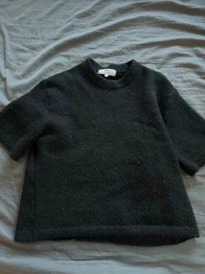 Svart stickad t-shirt från Gina Tricot - Svart stickad t-shirt från Gina Tricot i ullblandning. Tröjan har korta ärmar, rund halsringning och ribbstickade kanter. Perfekt för lager-på-lager och en stilren look.